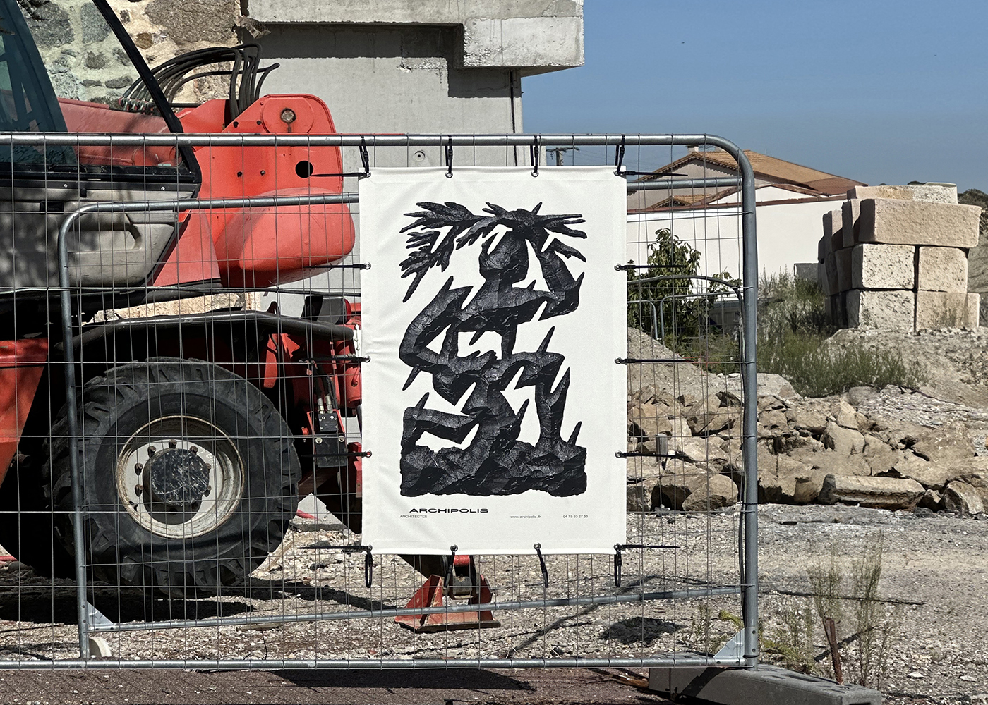 Photographie d'une bâche de chantier avec l'emblème d'Archipolis accrochée à une grille devant un tracteur rouge et une architecture en pierres.
