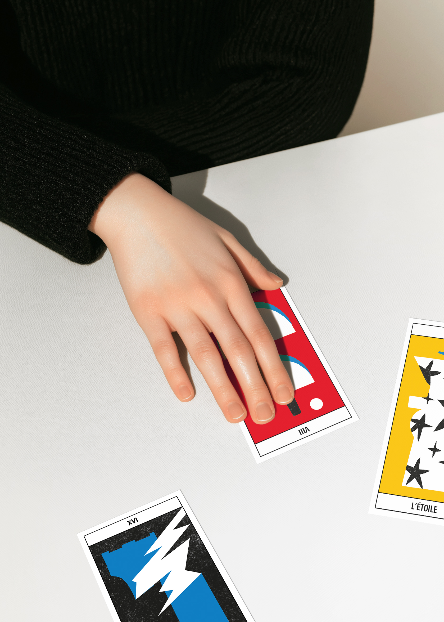 Main droite qui tire la carte Balance, numéro 8 du jeu de tarot Pompidou. Deux autres carte sont éparpillées sur la table, l'étoile et la tour.